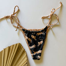Load image into Gallery viewer, A bikini bottom with side ties and a wild side animal print, displayed next to a golden palm-like object.
