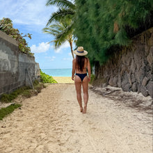 Load image into Gallery viewer, Woman walking along a sandy path towards the ocean with palm trees and a clear blue sky.